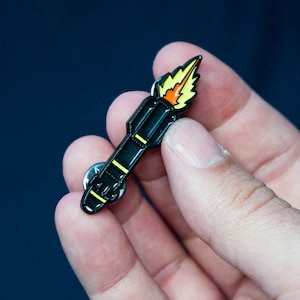 May include: Black enamel pin shaped like a rocket with a yellow and orange flame. The pin has a silver backing.