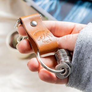 May include: A brown leather keychain with the text "Journey Made" embossed on it. The keychain has a metal ring and a set of metal key rings with the text "France" on one of the rings.
