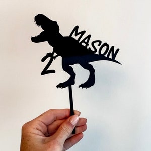 May include: Black silhouette of a dinosaur with the name Mason and the number 2 on a cake topper.