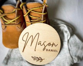 May include: A round wooden sign with the name "Mason Daniel" engraved in cursive script. The sign has a small leafy branch design below the name. The sign is sitting on a white onesie and brown baby shoes.