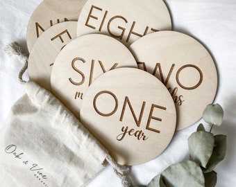 May include: A set of eight wooden baby milestone discs with engraved numbers and phrases, including "one year", "two months", "six months", and "eight months". The discs are in a white drawstring bag with the text "Oak & Vine Creations".