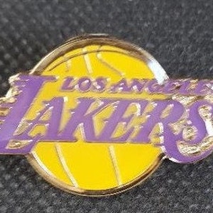 May include: A Los Angeles Lakers team logo pin. The pin features a yellow basketball with the team name in purple. The pin has a gold-colored border.