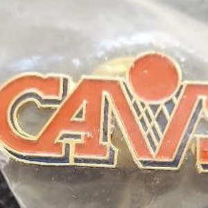 May include: A gold-colored pin with the word "CAVS" in red letters with black outlines. A basketball with black netting is behind the letters.