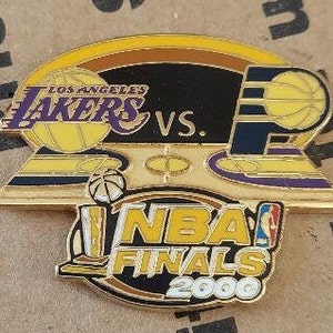 May include: A gold and purple NBA Finals 2000 pin featuring the Los Angeles Lakers logo and the text "VS." with a basketball court background.