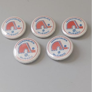 May include: Five round white buttons with a red and blue logo and the words "Nordiques Quebec". The logo features a stylized igloo shape. The buttons are arranged on a light gray surface.