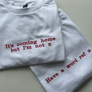 May include: Two white t-shirts with red embroidered text. The top shirt says "It's coming home but I'm not x". The bottom shirt says "Have a word ref x".