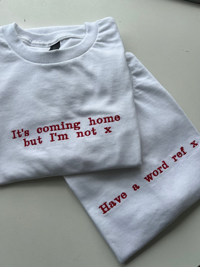 May include: Two white t-shirts with red embroidered text. The top shirt says "It's coming home but I'm not x". The bottom shirt says "Have a word ref x".