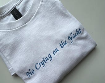 May include: White t-shirt with the embroidered phrase "No Crying on the Yacht" in blue script. The Gildan brand label is visible on the collar. The shirt is folded and lies on a light-colored surface.