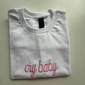 Puede incluir: Camiseta blanca con texto bordado en rosa que dice "cry baby"