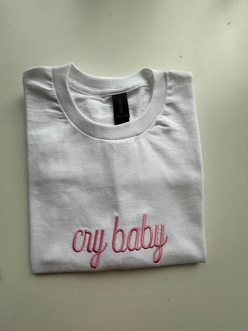 Puede incluir: Camiseta blanca con texto bordado en rosa que dice "cry baby"