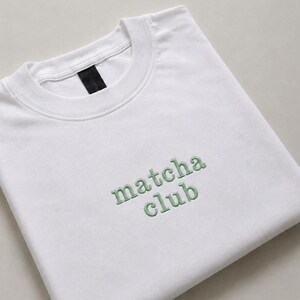 May include: A white crew neck t-shirt featuring the embroidered text "matcha club" in a light green thread centred on the chest. The garment includes a black interior neck label and has a ribbed collar design with visible stitching details.