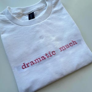 May include: White t-shirt with the phrase "dramatic much" embroidered in pink.