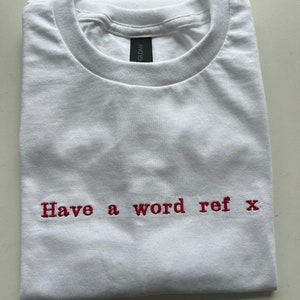 May include: White t-shirt with red embroidered text that reads "Have a word ref x".