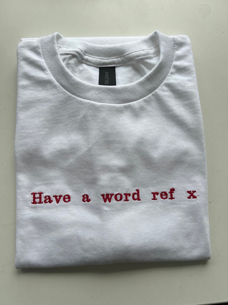 May include: White t-shirt with red embroidered text that reads "Have a word ref x".