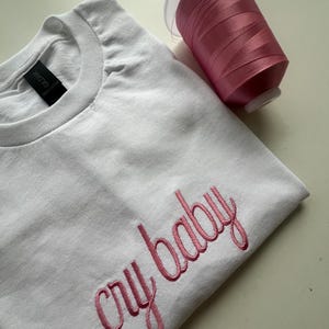 Puede incluir: Camiseta blanca con bordado rosa que dice "cry baby". Un carrete de hilo rosa est&aacute; al lado de la camiseta.