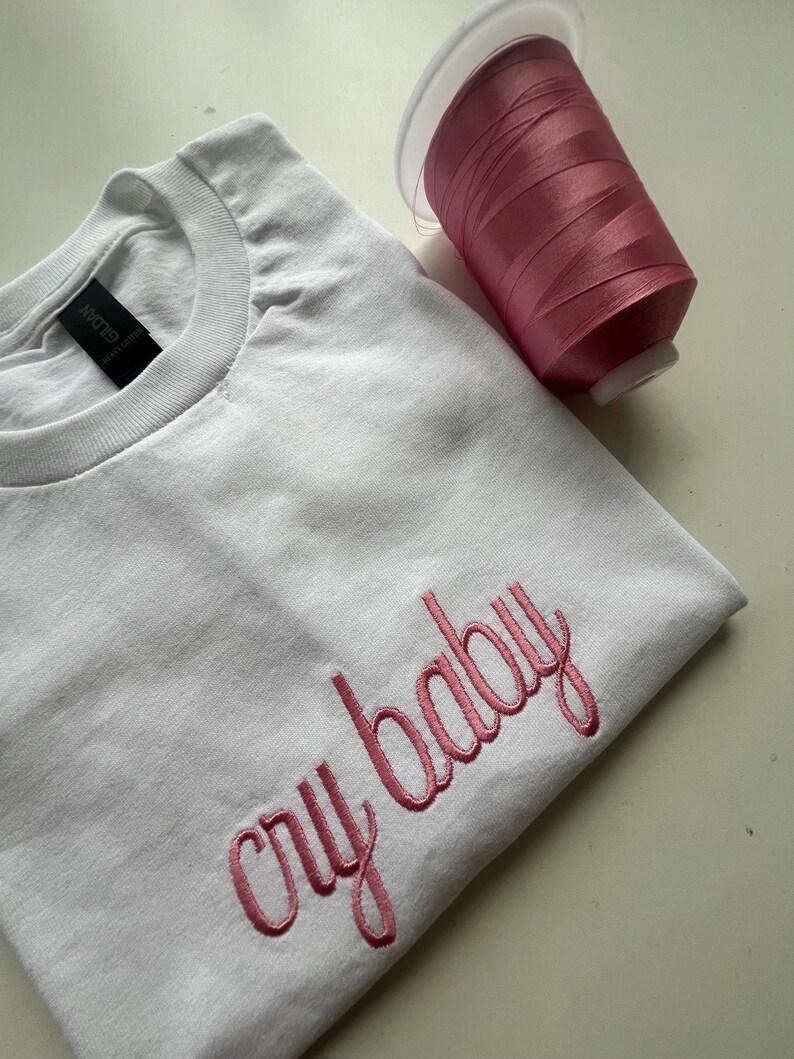 Puede incluir: Camiseta blanca con bordado rosa que dice "cry baby". Un carrete de hilo rosa est&aacute; al lado de la camiseta.