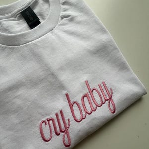 Puede incluir: Camiseta blanca con texto bordado en rosa que dice "cry baby"