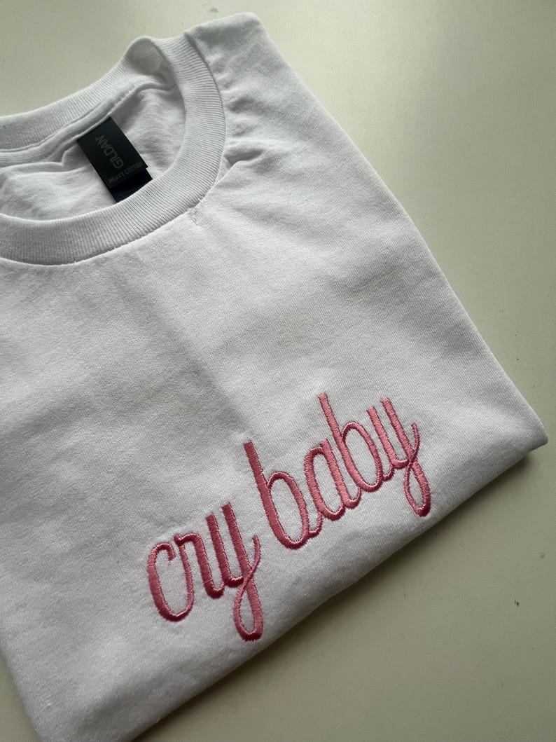 Puede incluir: Camiseta blanca con texto bordado en rosa que dice "cry baby"