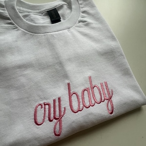 Puede incluir: Camiseta blanca con texto bordado en rosa que dice "cry baby"