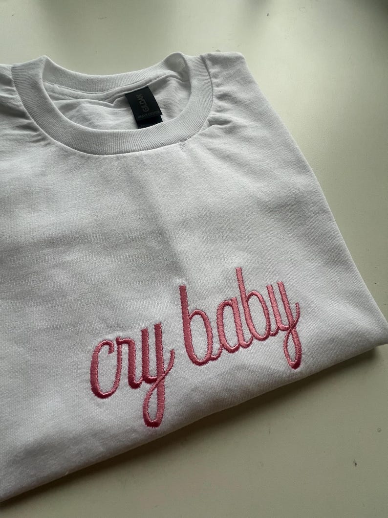 Puede incluir: Camiseta blanca con texto bordado en rosa que dice "cry baby"
