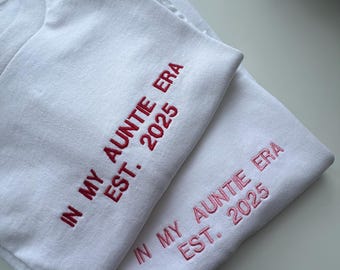 May include: Two white sweatshirts with the text "IN MY AUNTIE ERA EST. 2025" embroidered in red. The sweatshirts are folded and stacked on top of each other, showcasing the design.