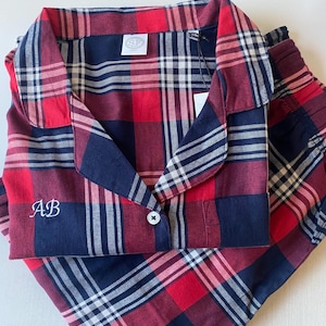Puede incluir: Conjunto de pijama a cuadros rojos, azul marino y blancos. La parte superior tiene cuello, cierre de botones e iniciales "AB" bordadas. El conjunto incluye una camisa de manga larga y pantalones a juego. Una etiqueta está adjunta.