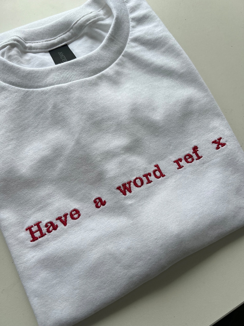 May include: White t-shirt with red embroidered text that reads "Have a word ref x".