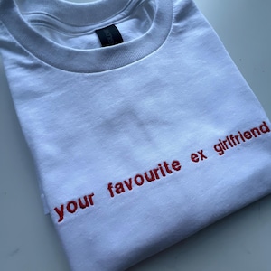 以下が含まれることがあります： 赤色の「your favourite ex girlfriend」という刺繍が施された、折りたたまれた白いTシャツ。クルーネックで、襟の近くに黒いタグが付いています。