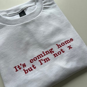 May include: White t-shirt with red embroidered text that reads "It's coming home but I'm not x"