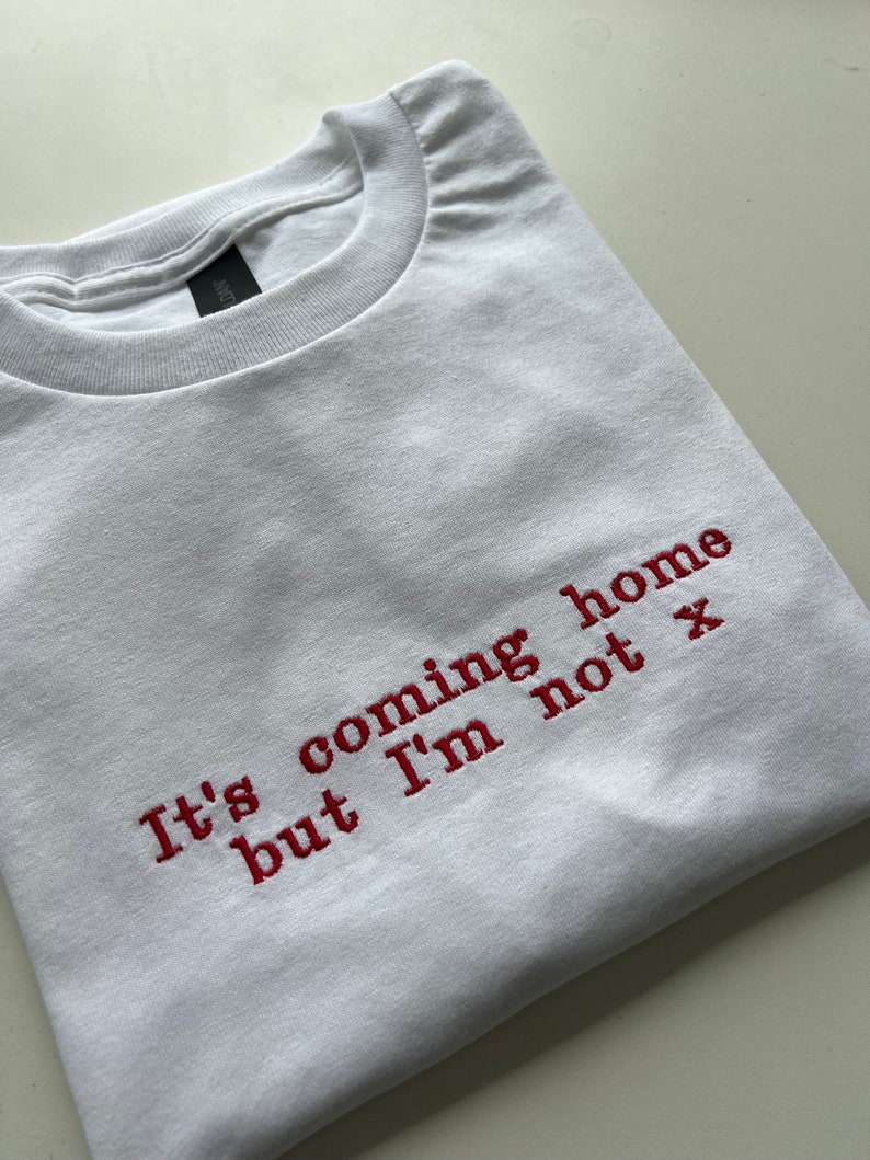 May include: White t-shirt with red embroidered text that reads "It's coming home but I'm not x"