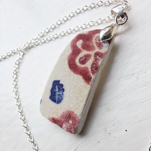 Puede incluir: Collar de cadena plateada con colgante de cerámica. El colgante es de cerámica blanquecina con diseños florales rojos y azules. El collar se muestra sobre una superficie blanca.