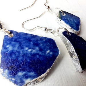 May include: A jewelry set featuring a necklace and earrings. The set includes a pendant necklace and two earrings, all crafted from irregular, dark blue ceramic pieces with silver-colored metal edges. The ceramic has a speckled white and blue pattern.