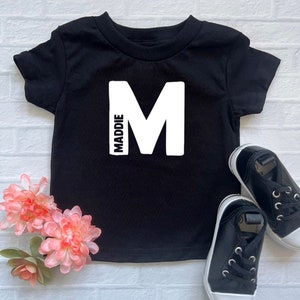 May include: Black short-sleeved t-shirt with a white letter "M" and the word "MADDIE" printed vertically on the left side of the shirt.