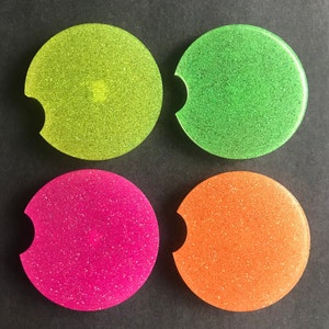 K&ouml;nnte beinhalten: Vier runde, glitzernde Untersetzer in Gr&uuml;n, Gelb, Pink und Orange. Die Untersetzer haben einen Ausschnitt f&uuml;r eine Tasse.