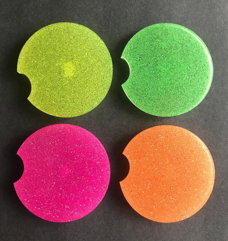K&ouml;nnte beinhalten: Vier runde, glitzernde Untersetzer in Gr&uuml;n, Gelb, Pink und Orange. Die Untersetzer haben einen Ausschnitt f&uuml;r eine Tasse.