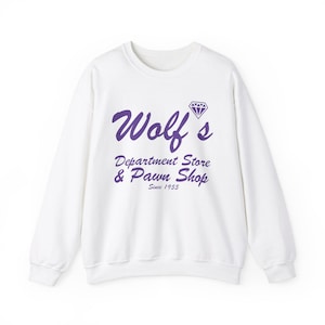 Könnte beinhalten: Weißer Pullover mit einer lilafarbenen Grafik, auf der "Wolf's Department Store & Pawn Shop Since 1955" steht, mit einem lilafarbenen Diamanten über dem Text.