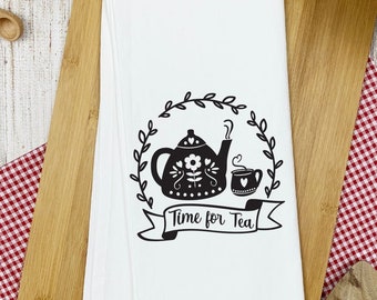 May include: White kitchen towel with a black graphic of a teapot, cup, and the text "Time for Tea." The towel is on a wooden cutting board, with wooden spoons and a red and white checkered cloth nearby.