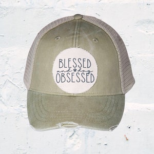 May include: Olive green distressed baseball cap with a mesh back. The front features a circular patch with the words "BLESSED and dog OBSESSED" in black script and a paw print. The cap has a pre-curved brim and a button on top.