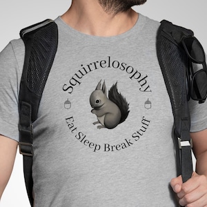 May include: A gray t-shirt featuring a circular design with a squirrel illustration and the words "Squirrelosophy Eat Sleep Break Stuff". The design includes two acorns. The shirt is made of a soft material.
