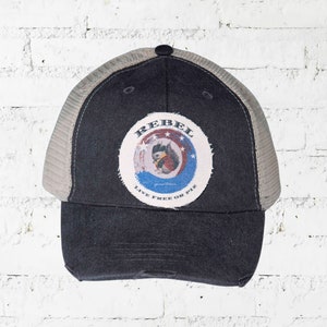 May include: A dark blue trucker hat with a distressed look. The hat features a circular patch with a rooster graphic and the word "REBEL". The patch is surrounded by a blue and red design with the text "LIVE FREE OR PIE".