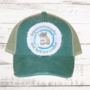 May include: A green trucker hat with a mesh back and a distressed brim. The hat features a circular patch with a squirrel wearing glasses and holding a coffee cup, with the text "Squirrelosophy not before coffee."