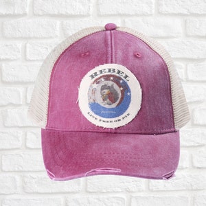 May include: A distressed magenta trucker hat with a white mesh back. The front features a circular patch with the word "REBEL" and an illustration. The text "LIVE FREE OR DIE" is below the illustration. The hat has a curved brim.