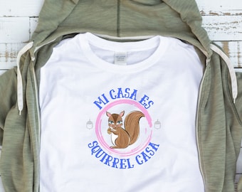 May include: White t-shirt with the text "Mi Casa Es Squirrel Casa" in blue and pink. The shirt features a cartoon squirrel holding a nut inside a pink circle. The shirt is displayed inside a green zip-up hoodie.
