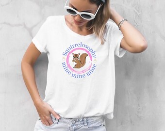 May include: White t-shirt featuring a cartoon squirrel design within a pink and blue circular frame. The text "Squirrelosophy" and "mine mine mine" are printed around the squirrel. The shirt is paired with denim shorts.