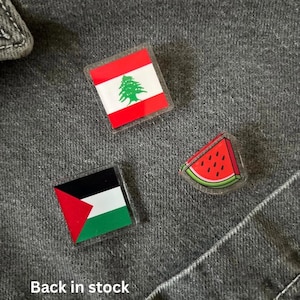 May include: Three pins on a grey denim background. The pins are clear plastic with images inside. The first pin is the flag of Lebanon, the second pin is the flag of Palestine, and the third pin is a slice of watermelon.