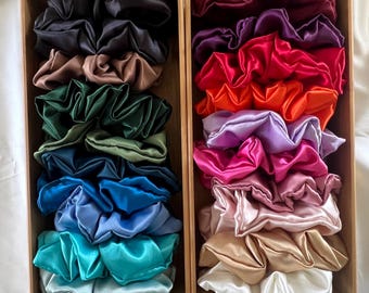 May include: Two wooden boxes filled with satin scrunchies in various colours. One box contains shades of black, brown, green, blue, and teal. The other box holds scrunchies in burgundy, purple, red, orange, pink, and white.