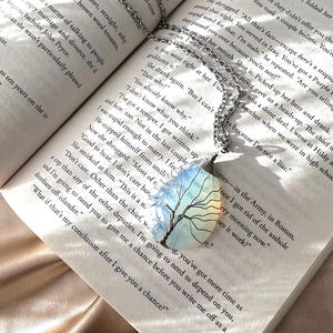 May include: A silver necklace with a pendant featuring a colourful tree design. The pendant is set against a page of a book with text about a person's military service.