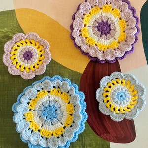 May include: Four crocheted coasters in different colors and patterns. The coasters are circular and have a flower-like design. The colors are purple, yellow, white, blue, and light blue.