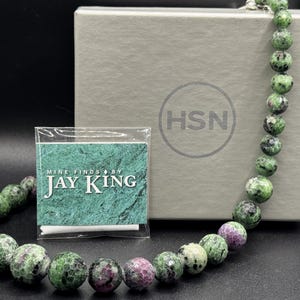 May include: A beaded necklace featuring round, polished stones in shades of mottled green and magenta. The necklace is displayed with a gray box marked with the HSN logo and a small square card labeled Mine Finds by Jay King in white text.