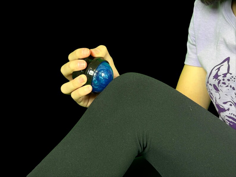 K&ouml;nnte beinhalten: Ein schwarzer und blauer Handtrainer mit strukturiertem blauem Innenraum. Der Trainer wird in einer Hand vor schwarzem Hintergrund gehalten. Der Trainer dient zur Verbesserung der Griffkraft und Fingerfertigkeit.